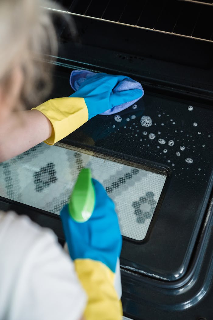 A person wearing gloves cleans an oven door using a spray and cloth.